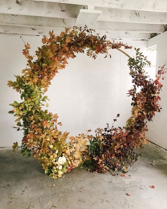 a round wedding arch covered with greenery, bold fall leaves and nothing else looks very organic and chic