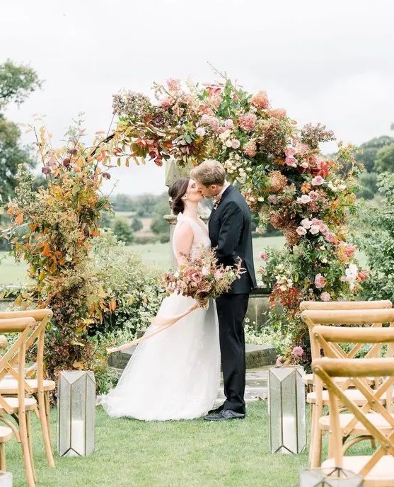 a round wedding arch with greenery and bold fall leaves, blush and pink blooms and some branches is a cool idea for a fall wedding