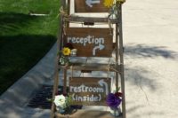 a rustic wedding decoration of a ladder, bright blooms and greenery and plywood signs is easy to DIY