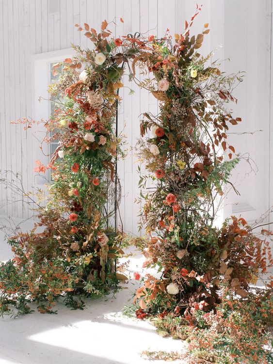 a stunning fall wedding arch decorated with greenery, branches, white and orange blooms looks wow