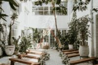 a stylish boho wedding ceremony space with stained benches, white blooms and greenery, a triangle wedding arch and lots of potted plants around