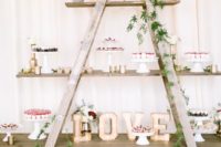 a stylish rustic wedding dessert bar of a ladder interwoven with greenery, with marquee lights and lots of delicious desserts