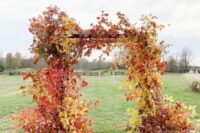 a super bold wedding arch completely covered with red and yellow fall leaves is a stunning idea for a bright fall wedding