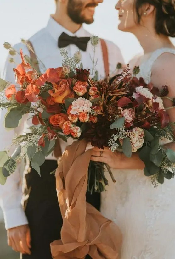 a super lush fall wedding bouquet of rust, blush, orange, burgundy blooms, lots of greenery and bright foliage, long tan ribbons is wow