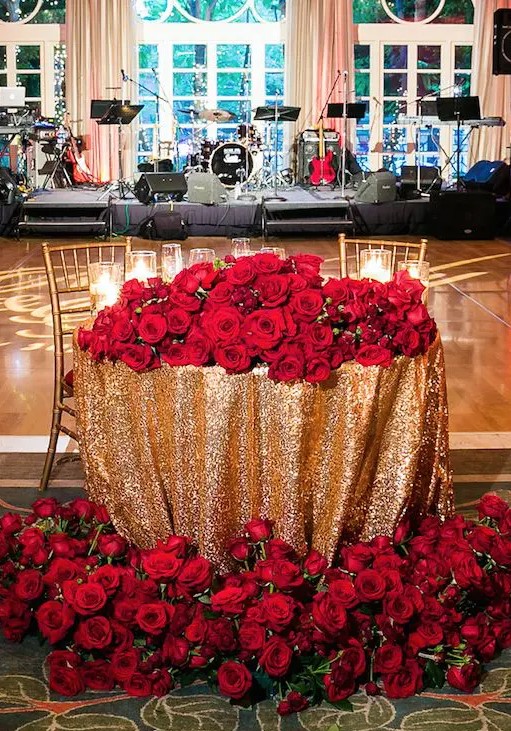 a sweetheart table with a gold sequin tablecloth and lots of red roses and greenery on the table and on the floor