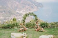 a unique wedding ceremony space with a sea view, a greenery and bloom wedding arch, kidney-shaped upholstered ottomans and pink and white blooms around them