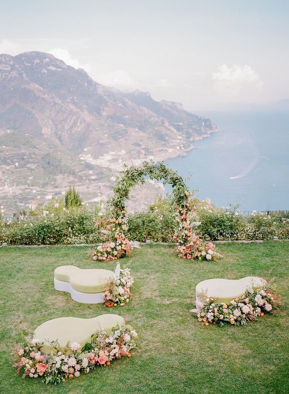 a unique wedding ceremony space with a sea view, a greenery and bloom wedding arch, kidney-shaped upholstered ottomans and pink and white blooms around them