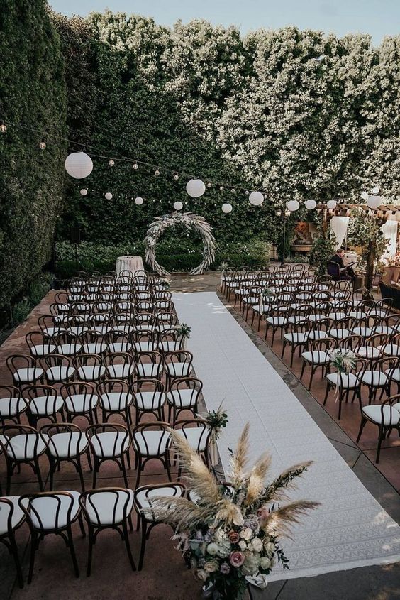 an epic boho wedding ceremony space with greenery walls, white chairs, pampas grass and blooms is a great idea for a neutral boho wedding