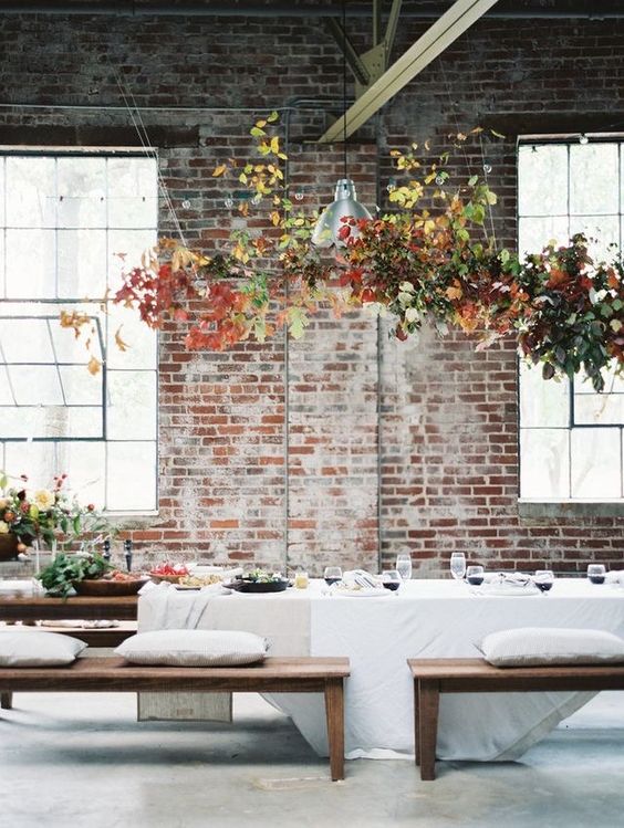 an industrial wedding reception space with greenery and bold leaves over the table is a cool room for a fall wedding