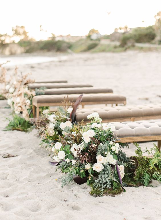 grey upholstered benches decorated with greenery, neutral and dark blooms are a stylish idea for a chic beach wedding