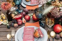 a beautiful fall harvest wedding tablescape with various fruit and berries including pomegranates on the table, burugndy candles, orange napkins, gold cutlery