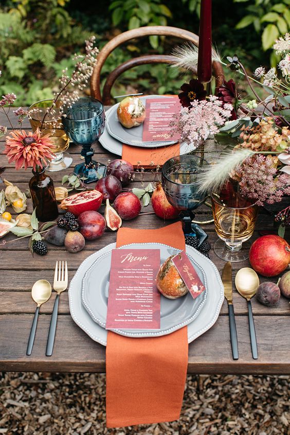 a beautiful fall harvest wedding tablescape with various fruit and berries including pomegranates on the table, burugndy candles, orange napkins, gold cutlery