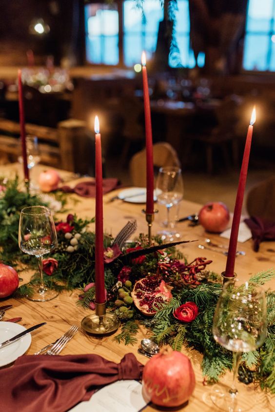 a boho winter wedding tablescape with an evergreen and red rose runner, feathers and burgundy candles, burgundy napkins and pomegranates
