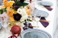 a bold and eye-catchy fall wedding tablescape with yellow, white and deep purple blooms, purple candles and napkins, pomegranates and gold cutlery