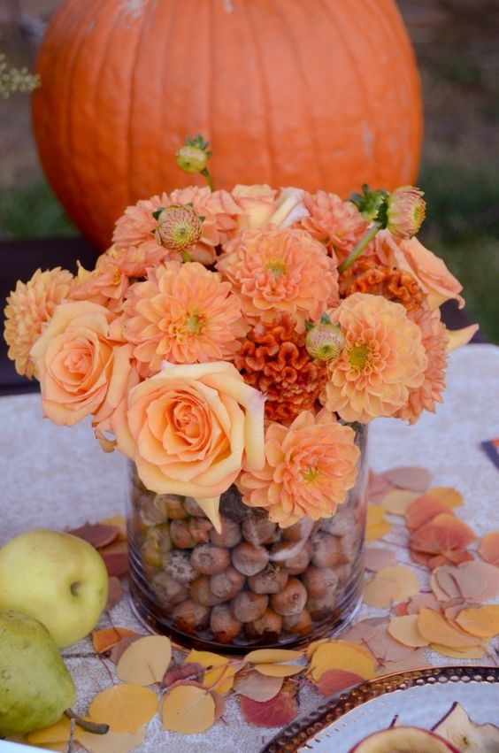 a bold fall wedding centerpiece of a glass filled with acorns, bold orange and rust blooms is a cool idea to go for