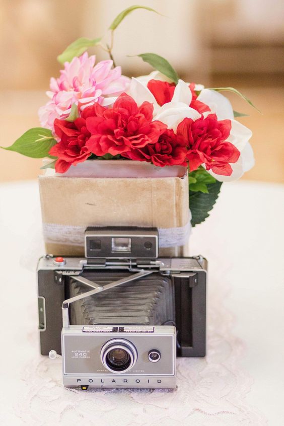 a catchy wedding centerpiece of vintage books, a vintage camera, bold blooms and greenery can be easily DIYed