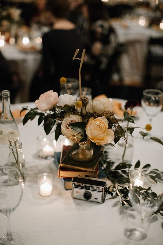 a chic wedding centerpiece of a stack of books, a beautiful floral wedding arrangement, a vintage camera and a gold table number