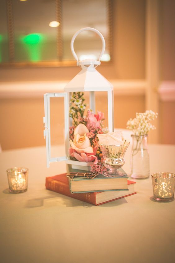 a couple of books, a glass with a white rose and a white lantern filled with fresh flowers for an elegant vintage centerpiece
