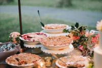 a cute summer wedding pie bar with pink blooms and greenery and pies on lovely stands or in plates is a cool idea to try