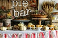 a gorgeous fall wedding pie bar with a colorful tassel garland, pinecones, white pumpkins, greenery, dried leaves and wheat