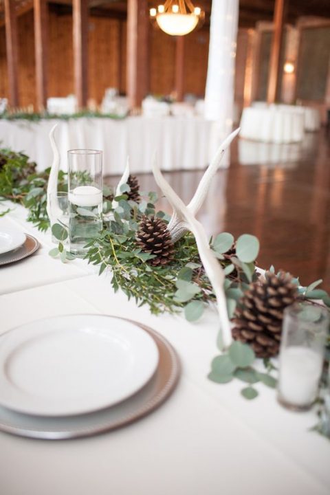 a greenery runner with pinecones, white candles and some antlers is a cool idea for a natural or woodland-inspired wedding