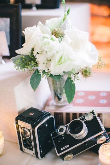 a lovely wedding centerpiece of a book, a white floral arrangement in a vase and a couple of vintage cameras is a stylish idea for a wedding