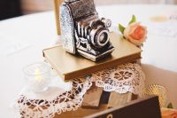 a lovely wedding centerpiece of a wooden crate with a doily, a book, a vintage camera and a table number plus candles around