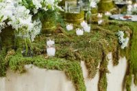 a lovely wedding tables covered with moss, with vases with white blooms wrapped with moss and lots of candles around for a woodland wedding