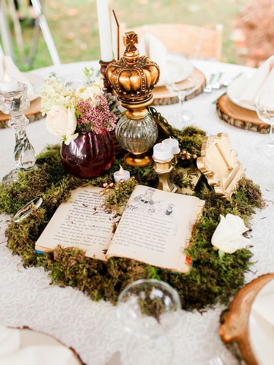 a magical wedding centerpiece composed of moss, an opened book, candles, vases with blooms and a table number