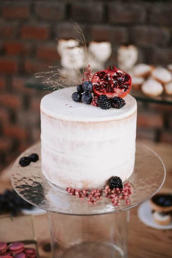 a naked wedding cake topped with blueberries, blackberries and pomegranates plus some grasses for a fall boho wedding