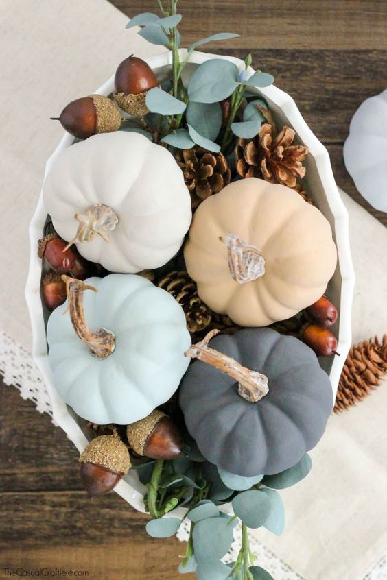 a pastel wedding centerpiece of a white bowl with pinecones, acorns, greenery and pastel pumpkins is a cool idea for the fall