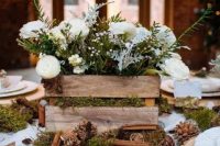 a rustic crate with greenery and flowers placed on moss with pinecones and cinnamon bark