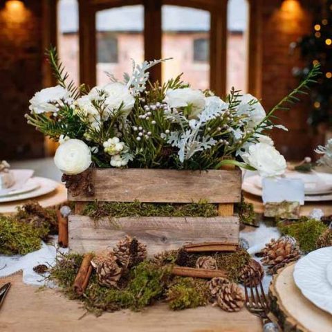 a rustic crate with greenery and flowers placed on moss with pinecones and cinnamon bark