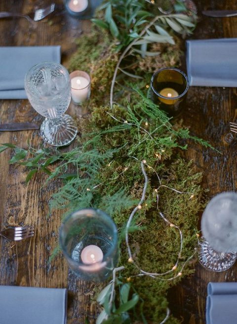 a simple uncovered wooden table with a moss and greenery runner, lights and candles plus grey napkins for a woodland wedding