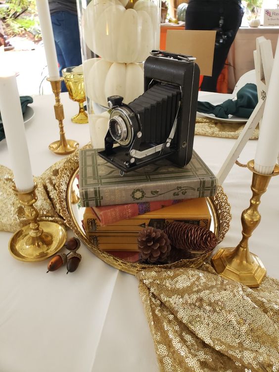 a unique fall wedding centerpiece of a tray with books, pinecones, a book stack, a camera and some candles around, a godl sequin table runner