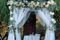 a wedding arch with white tulle curtains, lush greenery and neutral blooms is ideal for a rustic wedding