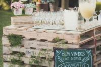 a wedding drink bar of pallets and ferns in between, with a chalkboard sign and floral arrangements on top
