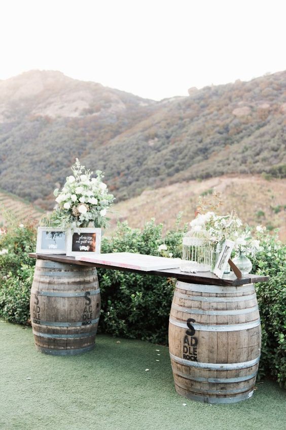 a wedding guest book placed on a stand of two wine barrels and a countertop, greenery, blooms and chalkboard signs in frames