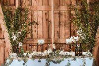 a wedding sweetheart table decorated with lots of greeneyr and white blooms plus long branches behind the table as a natural backdrop