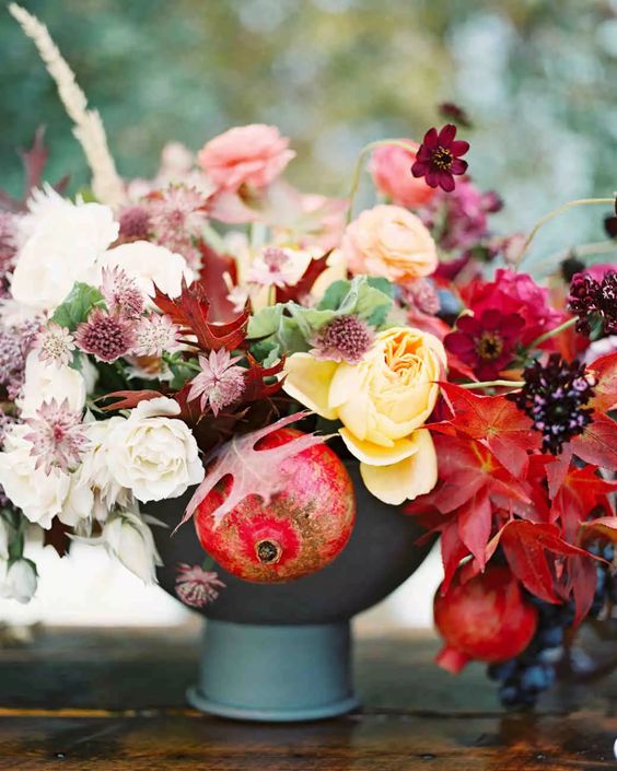 an adorable fall wedding centerpiece of a grey vase, yellow, pink and deep red blooms, fall leaves and pomegranates that help to embrace the fall