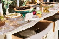 an elegant wedding pie bar of a vintage sideboard, with plates and cutlery in drawers, some blooms and greenery, fruits and delicious pies