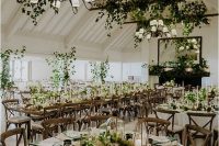 greenery arrangements on the wooden beams, chandeliers and tables make this indoor wedding recpetion space feel like an outdoor one