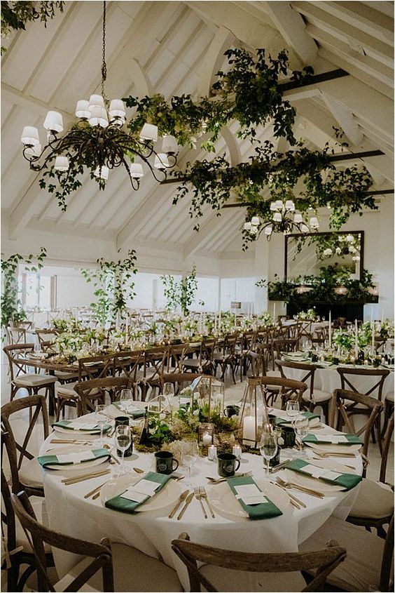 greenery arrangements on the wooden beams, chandeliers and tables make this indoor wedding recpetion space feel like an outdoor one