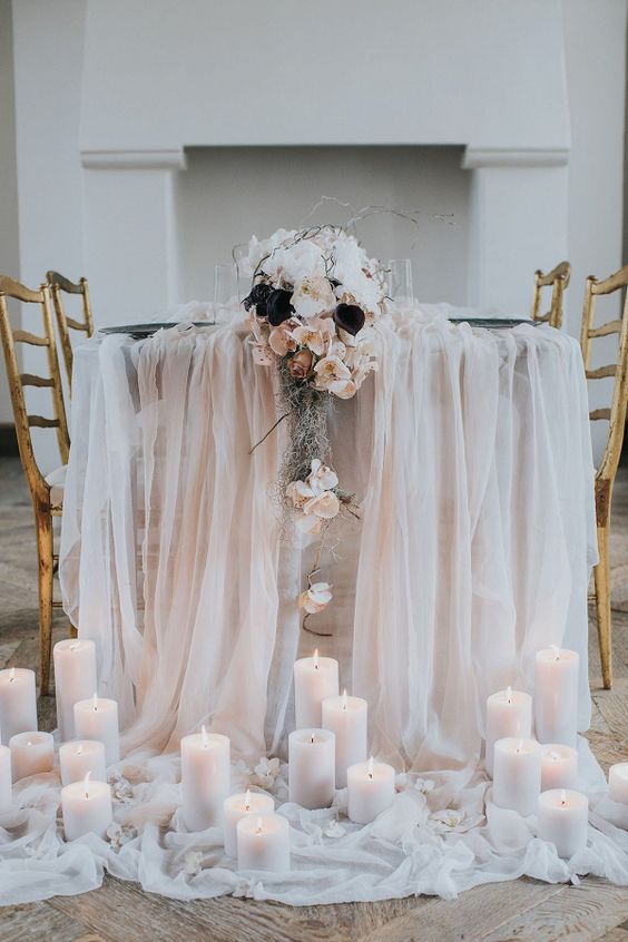 Изысканный декор стола для приема гостей с белым тюлем, свечами на столбиках и каскадным цветочным украшением