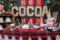 a Christmas hot cocoa bar with paper and pompom garlands, mugs, sweets and candies and a plaid tablecloth