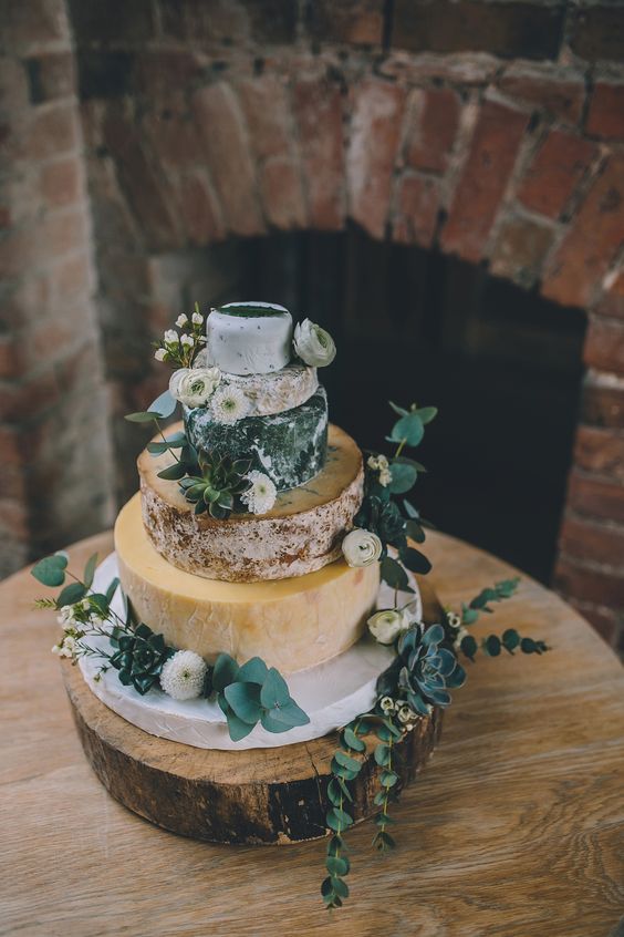 a beautiful cheese wheel wedding cake topped with eucalyptus and white blooms and displayed on a wood slice for a rustic wedding