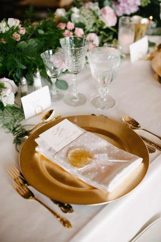 a beautiful neutral wedding tablescape with lots of greenery, blush, white and lilac blooms, a gold charger and cutlery, a gold lollipop as a favor