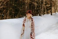 a boho winter bride wearing a boho lace sheath wedding dress, a burgundy hat and a printed coverup in burgundy and white