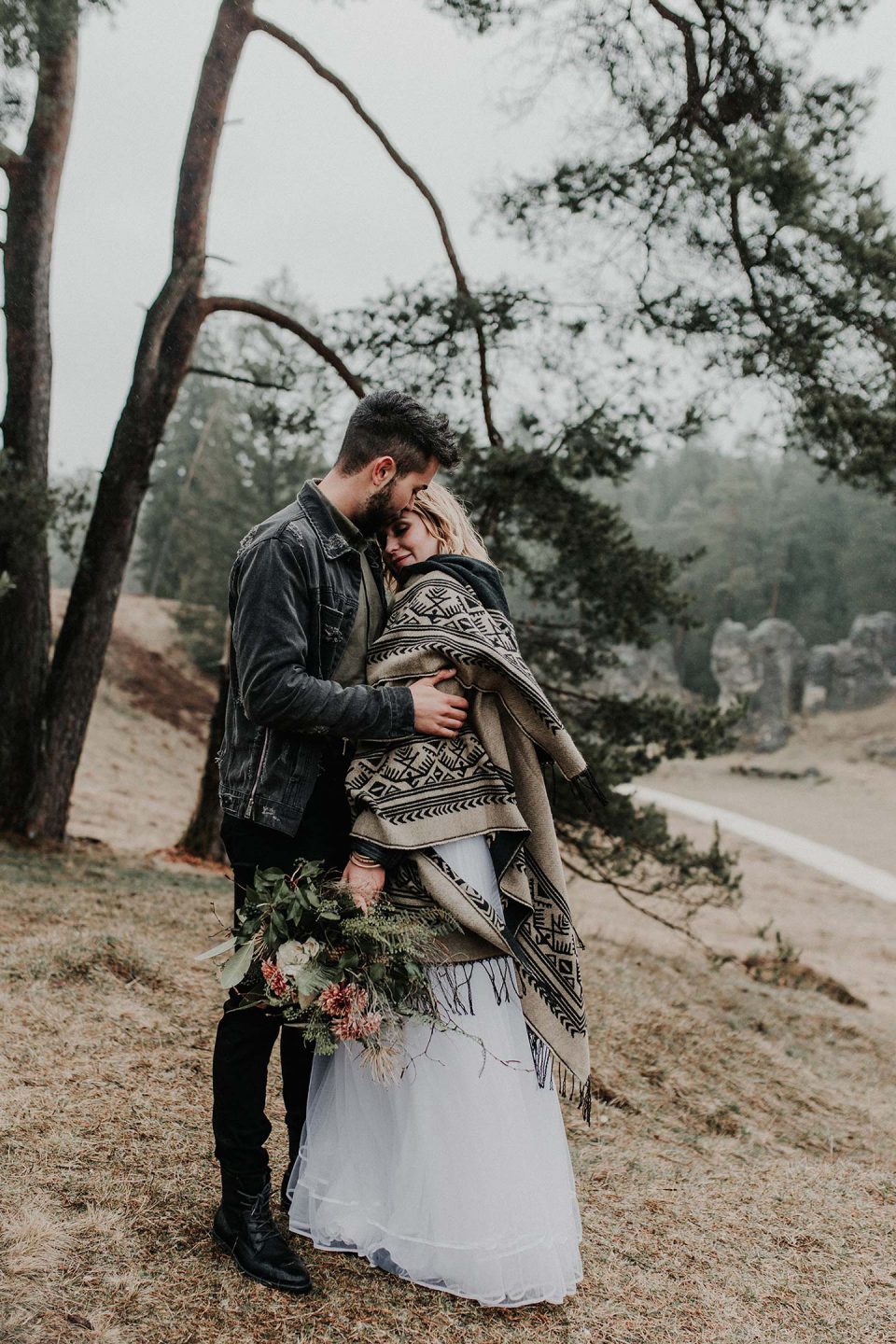 A bride covered up with a boho fringed printed piece and a groom wearing a black leather jacket and boots