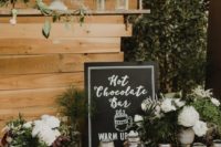 a chic and natural hot chocolate bar with a sign, jars with candies and toppings, mugs, greenery and neutral blooms